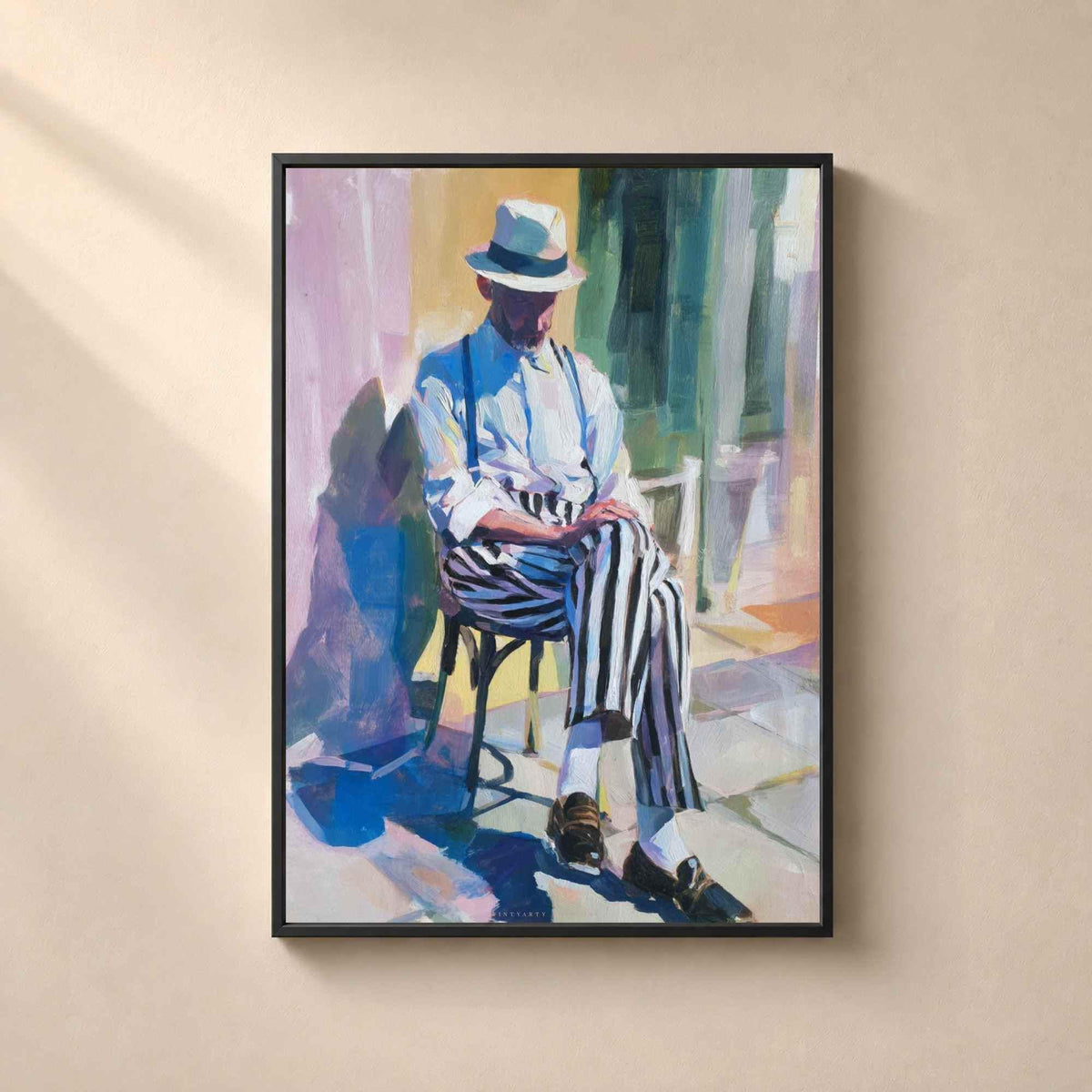 Affiche dans un cadre en métal noir fin sur un mur beige, homme assis au chapeau panama et pantalon rayé dans une rue méditerranéenne ensoleillée.