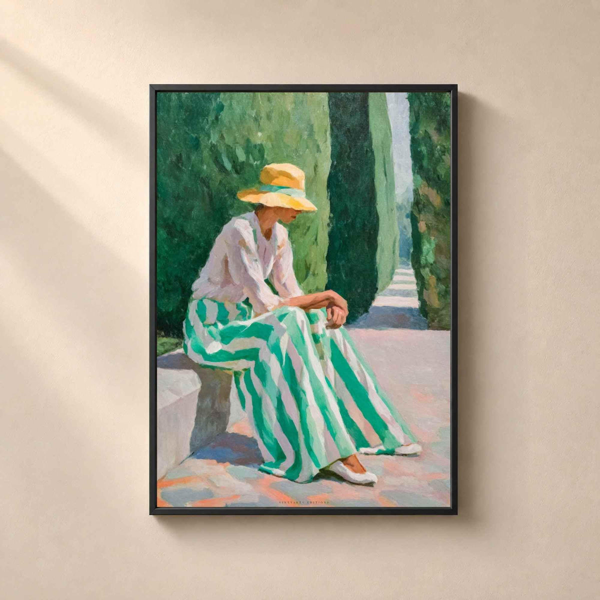 Affiche dans un cadre en métal noir fin sur un mur beige, femme élégante au chapeau de paille et jupe rayée verte assise dans une allée de cyprès.