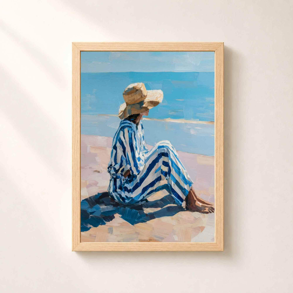 Affiche dans un cadre en chêne naturel sur un mur clair, femme assise de profil sur le sable en tenue rayée bleue et chapeau de paille face à la mer.