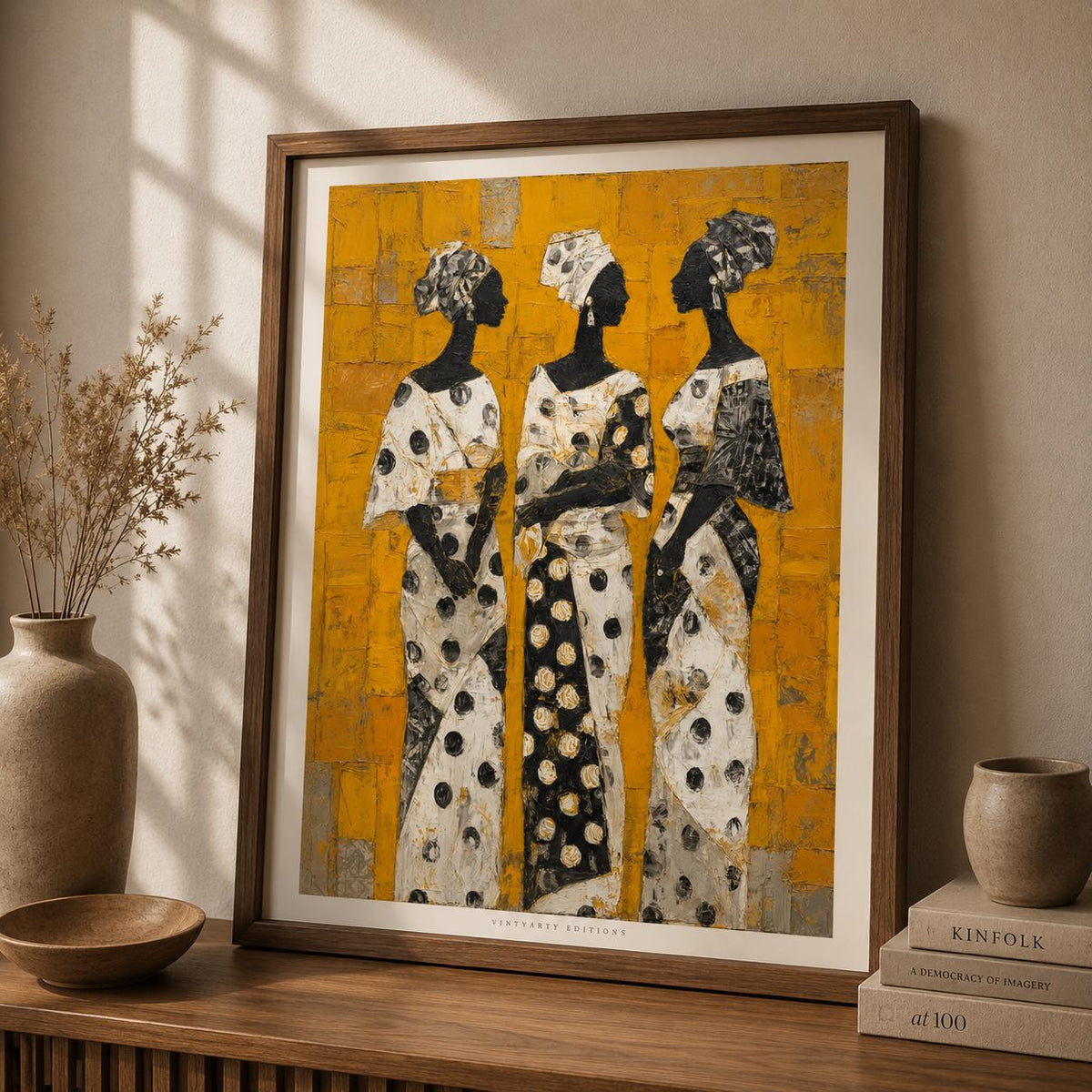 Affiche trois femmes africaines dans un cadre en bois sombre sur un mur clair, trois femmes en boubous à pois noir et blanc sur fond jaune d'or texturé.