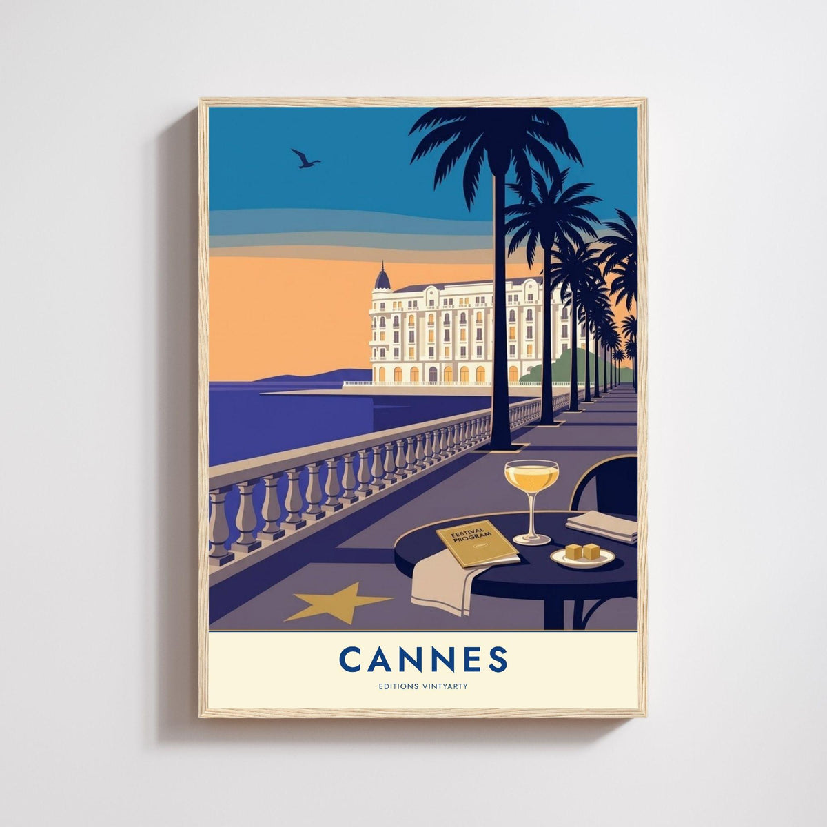 Affiche Cannes dans un cadre en chêne naturel sur un mur clair, Croisette au crépuscule avec palmiers, hôtel blanc et table dressée face à la mer.