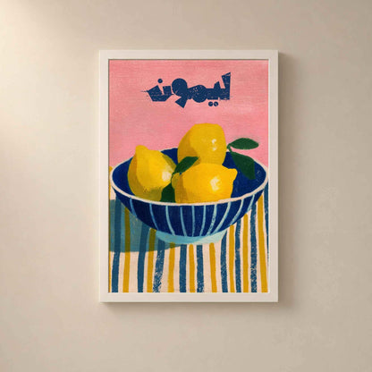 Affiche citrons dans un cadre en bois blanc sur un mur clair, trois citrons dans un bol bleu rayé sur une nappe jaune et bleue, fond rose.