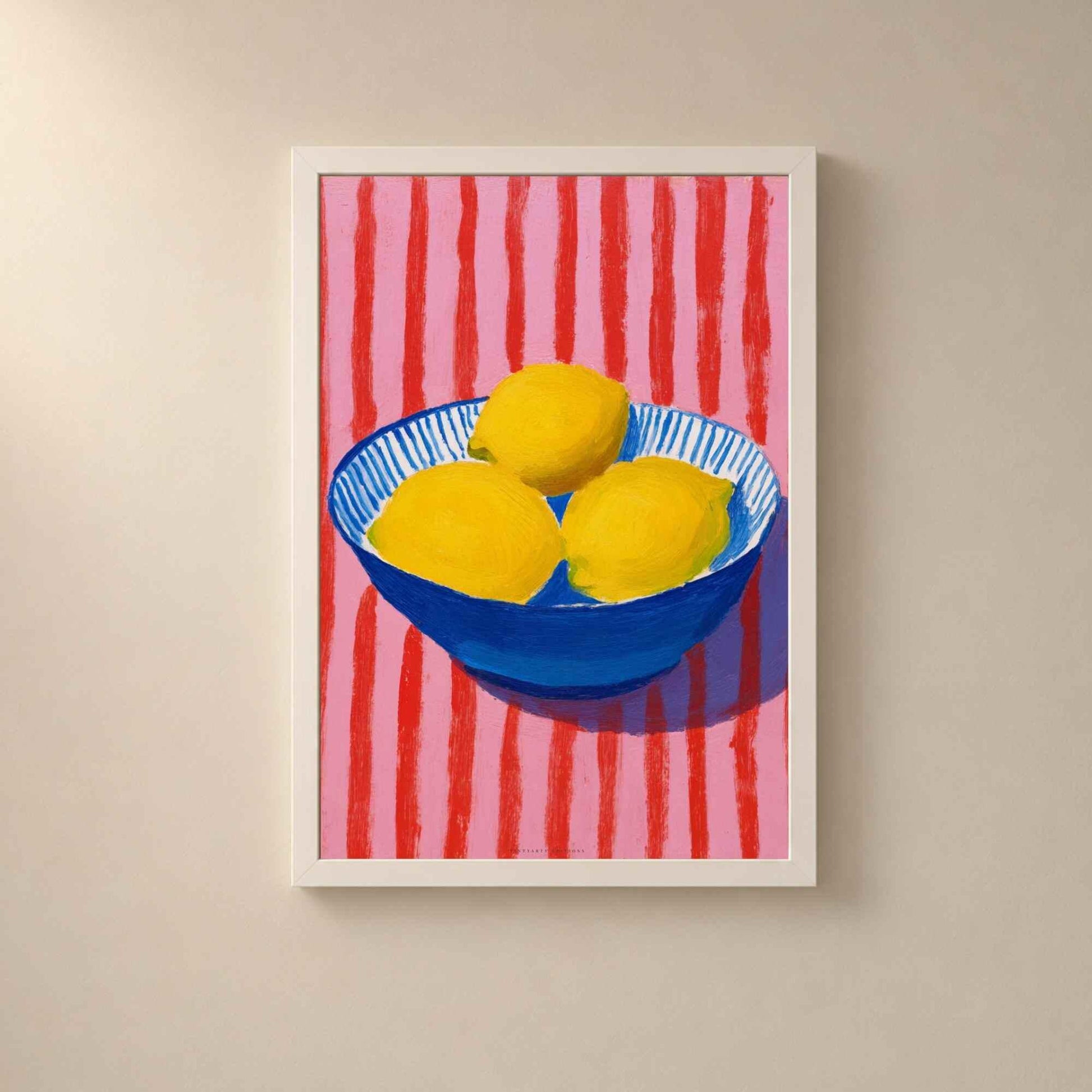 Affiche citrons dans un cadre en bois blanc sur un mur clair, trois citrons dans un bol bleu sur un fond rose rayé de rouge.