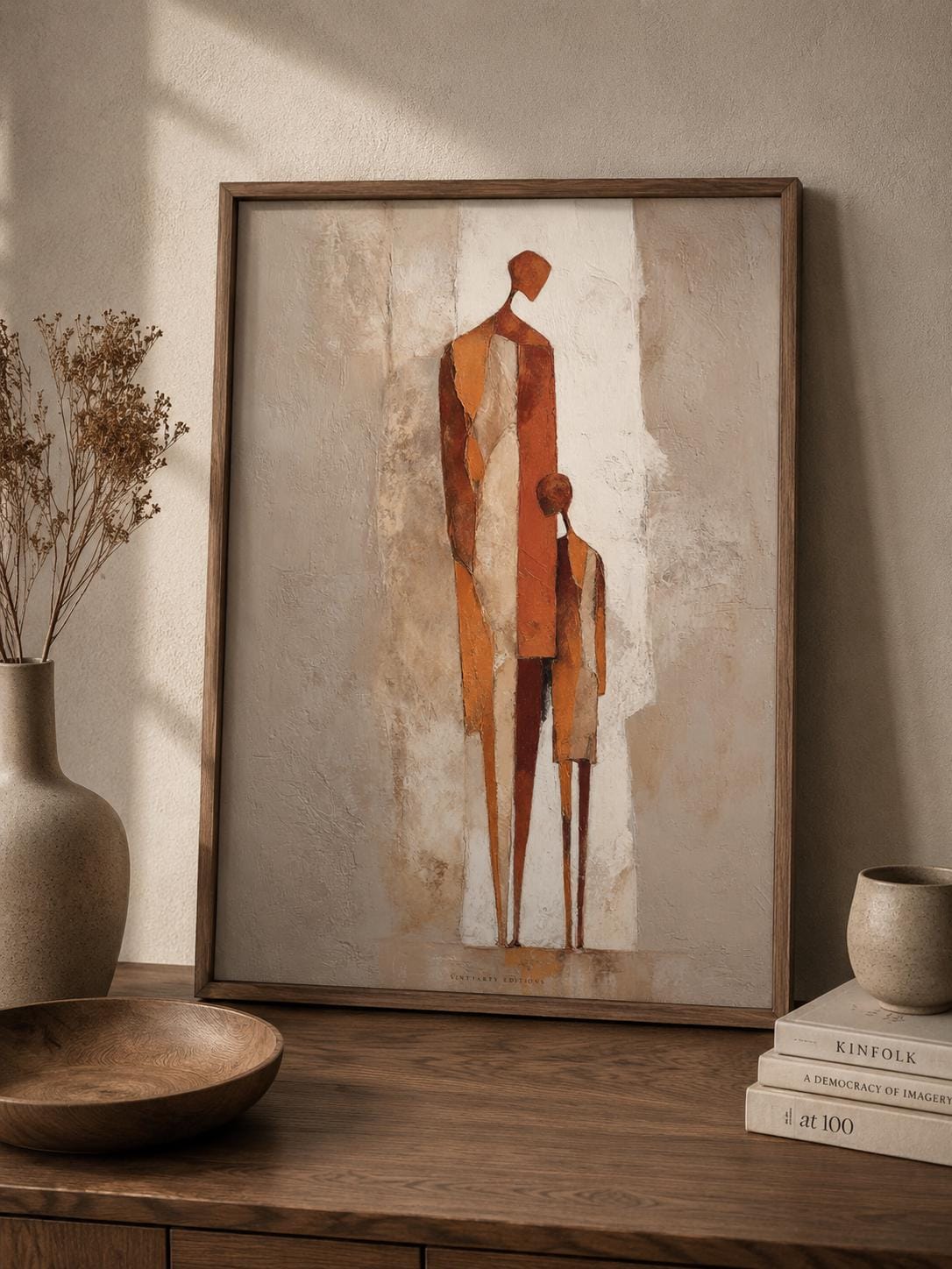 Affiche parent et enfant dans un cadre en bois sombre sur un mur clair, silhouettes abstraites drapées de terracotta et rouille sur fond patiné.