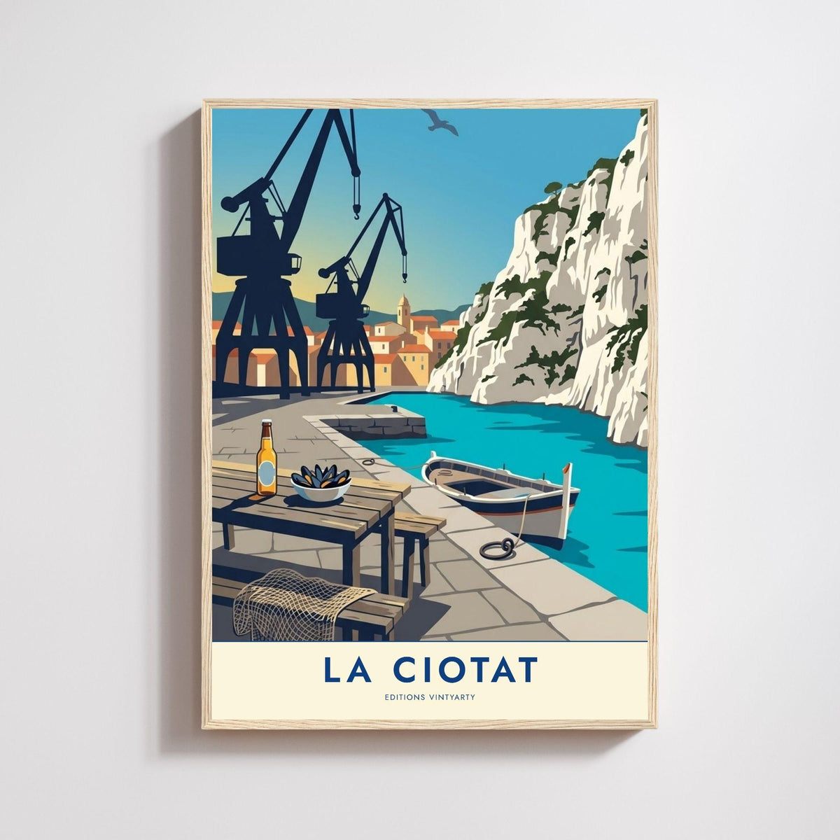 Affiche La Ciotat dans un cadre en chêne naturel sur un mur clair, grues de chantier naval noires face à une falaise blanche et une barque turquoise.